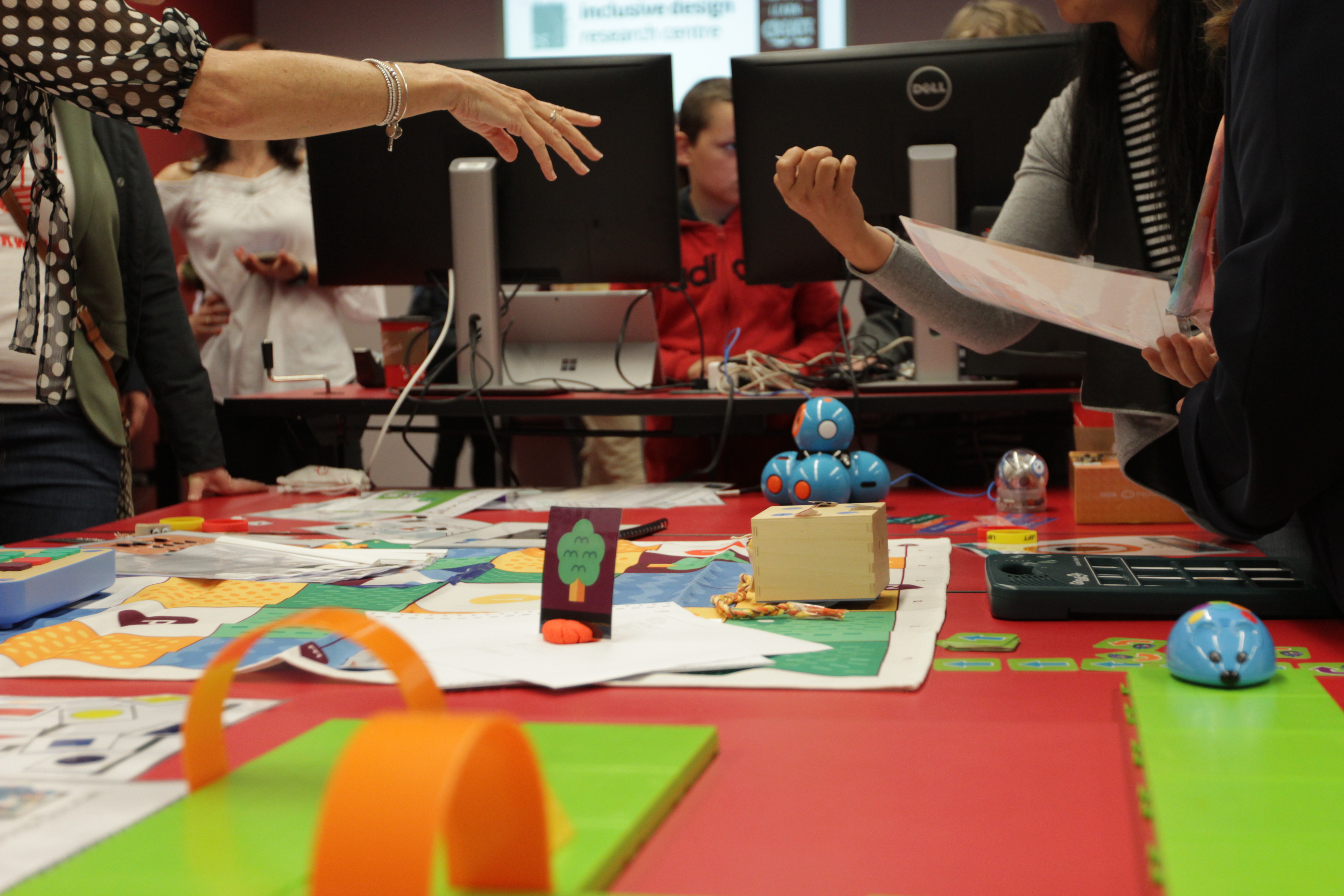 Several people having a conversation around a table with coding toys and robots on it.